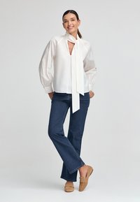 Blusa bianca con scollatura a V e dettaglio con fiocco, dotata di maniche a sbuffo; abbinata a jeans svasati blu scuro e mocassini beige.