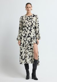 Vestido floral negro y crema con mangas largas, cuerpo plisado, dobladillo con volantes y abertura hasta el muslo, combinado con botas negras hasta la rodilla.