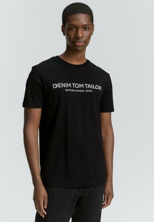 Sort T-shirt med korte ærmer lavet af bomuld og med rund halsudskæring. Har en hvid logo-tekst "DENIM TOM TAILOR ETABLEREDE 2007" hen over forsiden.