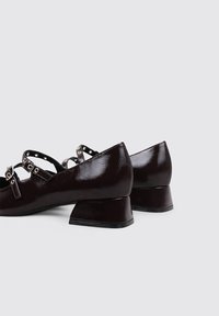 Zapatos de charol marrón con un tacón triangular bajo, que cuentan con correas ajustables con herrajes de metal y un acabado texturizado.