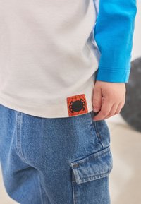 Maglietta bianca con maniche lunghe blu; presenta una toppa con logo di un ragno rosso sull'orlo. Indossata con pantaloni cargo di jeans blu.