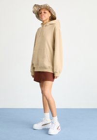 Beżowy hoodie z kieszenią z przodu, brązowe szorty i masywne białe sneakersy. Uzupełnione leopardskim kapeluszem bucket, stojące na niebieskim tle.