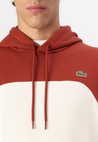 Sudadera de algodón roja y crema con un bolsillo tipo canguro, cordones y un pequeño logo bordado en el pecho. Textura suave con colores contrastantes.