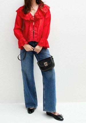 Donna che indossa una blusa rossa in pizzo a occhiello con colletto arricciato, jeans a gamba larga blu, ballerine nere e tiene una piccola borsa nera.