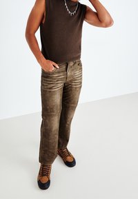 Brun ärmlös topp, slitna olivgröna byxor med vertikala linjer, och bruna vandringsskor med svarta sulor och snören. Enfärgad bakgrund.