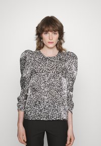 Mulher com cabelo castanho na altura dos ombros veste blusa de mangas balão em preto e branco e calças pretas, de pé contra um fundo simples.