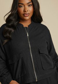 Svart zip-up jacka med rund halsringning, två framfickor och texturerat tyg. Har elastiska ärmslut och en avslappnad passform.