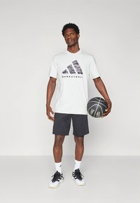 Camiseta blanca con logo de Adidas en negro y morado, combinada con pantalones cortos negros, sosteniendo un balón de baloncesto, con calcetas blancas y zapatillas deportivas negras.