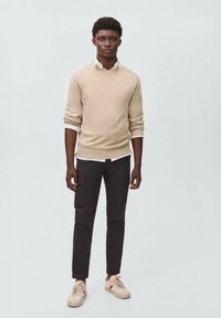 Bež pleteni pulover preko bele srajce s ovratnikom, v kombinaciji s temnimi cargo hlačami in bež supergami, postavljen na nevtralni ozadju.