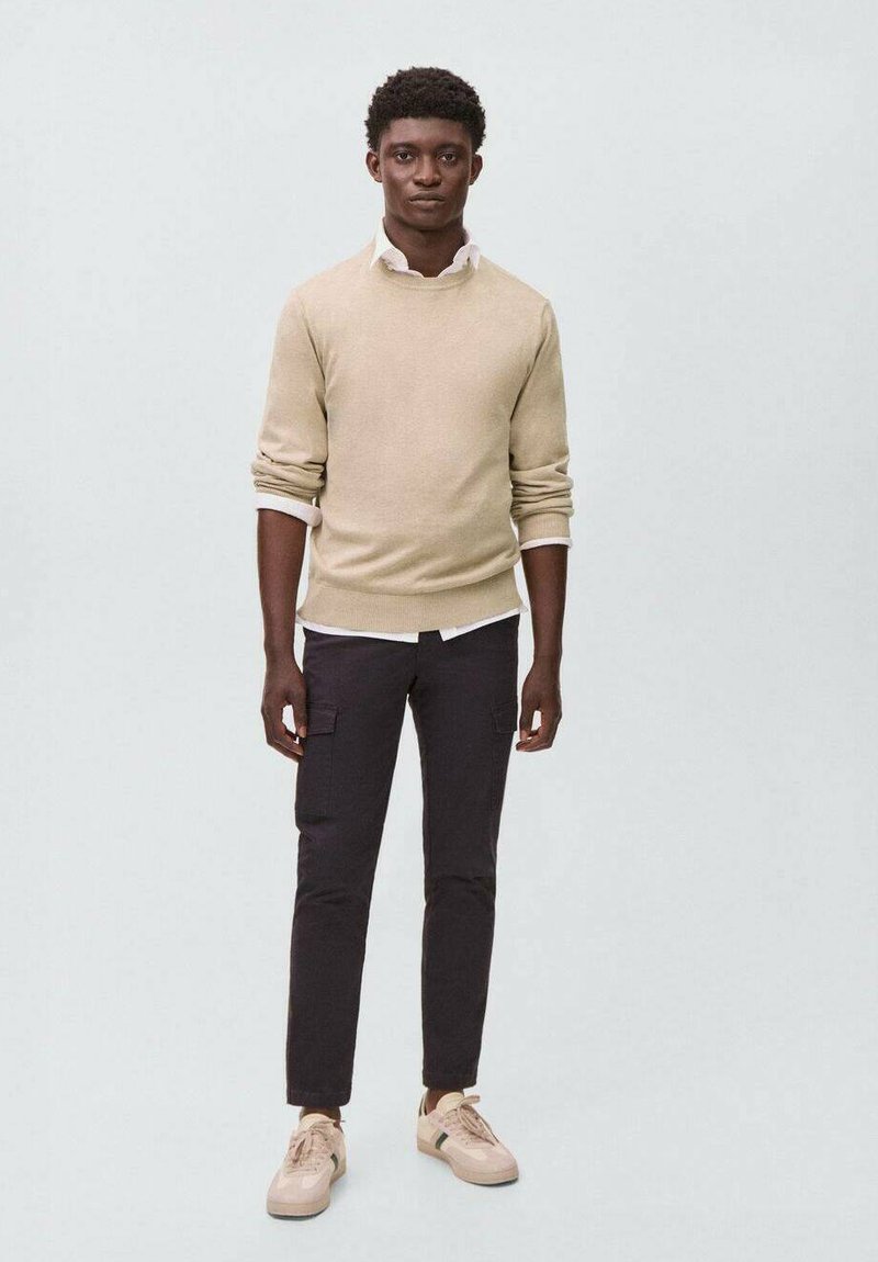 Bež pleteni pulover preko bele srajce s ovratnikom, v kombinaciji s temnimi cargo hlačami in bež supergami, postavljen na nevtralni ozadju.