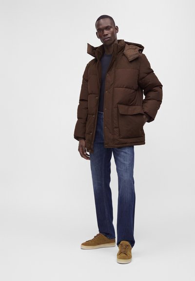 Homme portant une doudoune marron foncé, un jean bleu foncé et des baskets marron clair, debout avec une main dans la poche sur fond blanc.