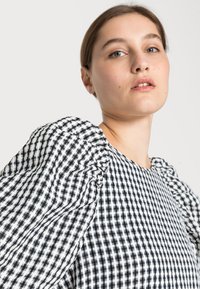 Blusa xadrez preto e branco com mangas bufantes, apresentando um padrão texturizado e um tecido liso, inclinada para cima para uma vista em detalhe.