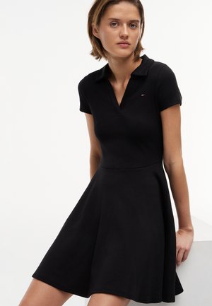 Mladá žena oblečená v krátkorukávových čiernych šatách typu polo s priliehavým pásom a rozšírenou sukňou, sediaca s neutrálnym výrazom.