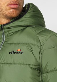 Zelená péřová bunda s kapucí, která má viditelné logo v černé a červené barvě. Materiál působí hladce a leskle a má prošívané části.