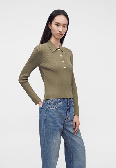 Femme aux longs cheveux noirs portant un polo côtelé à manches longues vert olive et un jean bleu large, debout, une main dans la poche.