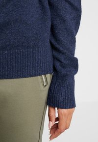 Pull en tricot bleu marine avec poignets côtelés, associé à un pantalon vert olive doté d'une poche zippée. Tissu texturé visible.