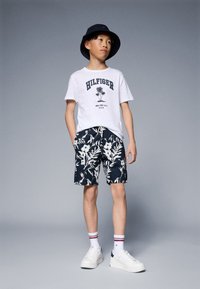 Hvit T-skjorte med blå "HILFIGER" tekst, svarte blomstrete shorts, hvite sneakers og en marineblå bøttehatt. Modellen står mot en grå bakgrunn.