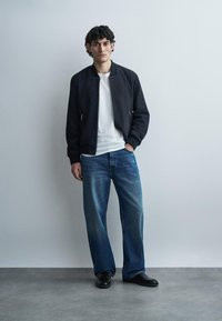 Vīrietis stāv pie vienkāršas sienas, valkājot melnu jaku, baltu T-kreklu, vaļīgas zilas džinsas un melnas kurpes, ar vienu roku kabatā.