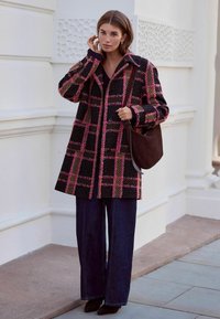 Schwarzer und rosa kariertes Mäntel mit großen Knöpfen, breiten Ärmeln und einer lässigen Passform, kombiniert mit dunkelblauen weit geschnittenen Jeans und einer braunen Schultertasche.