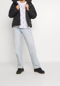 Czarna pikowana kurtka puchowa, biały t-shirt z fioletowym wzorem ananasa, jasnoniebieskie jeansy o prostych nogawkach oraz czarne buty z grubą podeszwą.