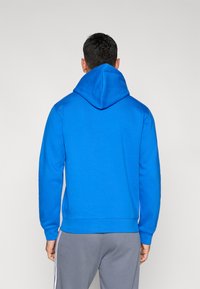 Mikina s kapucňou v v�ýraznej modrej farbe, vyrobená z jemnej látky. Má dlhé rukávy a klasický strih, s viditeľnými švami na prednej a zadnej strane.