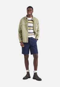 Camisa verde de botones sobre un suéter a rayas, pantalones cortos azul marino y botas negras. El modelo está de pie frente a un fondo blanco.