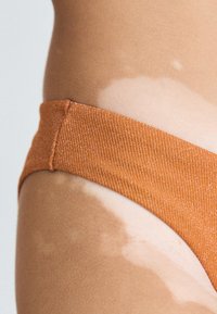 Narandžaste teksturirane gaćice s rebrastim dizajnom i suptilnim sjajem, svedene na srednji struk, koje prikazuju svijetle tonove kože s različitim pigmentacijama.