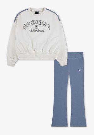Sweat blanc Converse avec bandes bleues sur les épaules et logo, porté avec un pantalon flare bleu avec un petit logo Converse sur la jambe gauche.