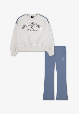 Sweat blanc Converse avec bandes bleues sur les épaules et logo, porté avec un pantalon flare bleu avec un petit logo Converse sur la jambe gauche.