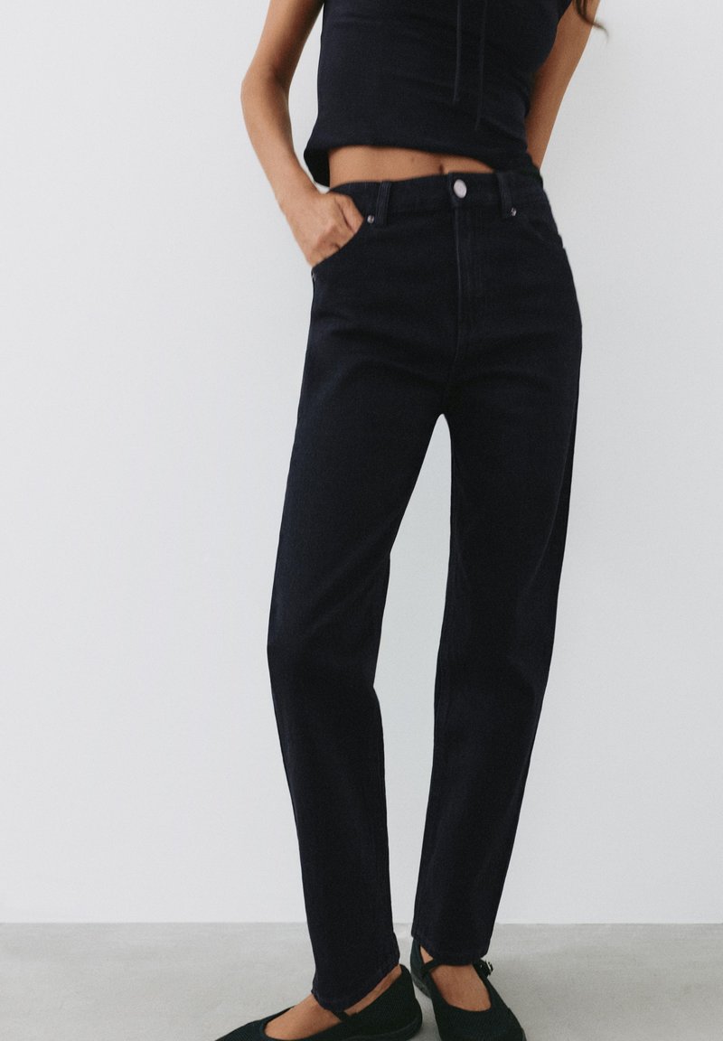 Femme portant un jean noir taille haute et un top noir court sans manches, debout avec une main dans la poche et des chaussures plates noires.