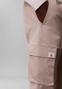 Primer plano de pantalones cargo beige con un gran bolsillo lateral y un parche visible con el logo de Calvin Klein cerca de la solapa del bolsillo.