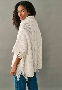 Gestrickter Poncho in Creme mit hohem Kragen, versehen mit Zopfmuster und fransigen Kanten. Kombiniert mit lockeren blauen Jeans.