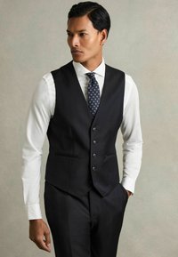Vesta neagră, croit�ă, dintr-un material fin, cu șase nasturi, două buzunare și un design ajustat, purtată cu o cămașă albă și o cravată cu model.