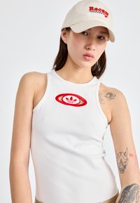 Balta šonkaite atrodančio topiuko su raudona išsiuvinėta "Adidas" logotipu, derinama su šviesiai beige skrybėle, ant kurios užrašytas raudonas žodis "Recap".