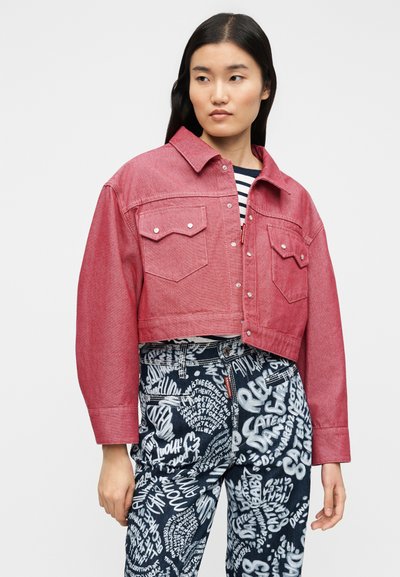 Veste courte en denim rouge avec deux poches poitrine, fermetures à boutons et finition texturée, associée à un jean bleu avec texte graphique blanc.