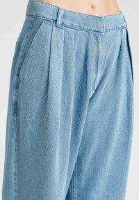 Pantaloni in denim azzurro chiaro con pieghe, caratterizzati da punti testurizzati su tutto il tessuto. Include tasche laterali e un design classico della vita.
