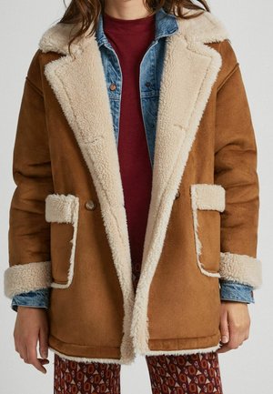 Mujer con un abrigo de piel con pelo interior color beige y ribete de piel color crema sobre una chaqueta vaquera azul y una camiseta color burdeos, de pie contra un fondo sencillo.