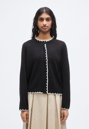 Μαύρο πλεκτό ζακέτα με λευκή κυματιστή διακόσμηση και κουμπιά κατά μήκος του μπροστινού μέρους, με μακριά μανίκια και στρογγυλή λαιμόκοψη.