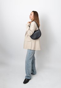 Femme en blazer beige et jean clair, debout de profil, tenant un sac à bandoulière en cuir noir sur un fond clair uni.