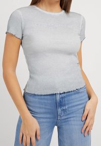 Top corto de canalé gris claro con mangas cortas, que presenta un dobladillo scalloped y un logotipo bordado en el frontal. Combinado con unos jeans azules.