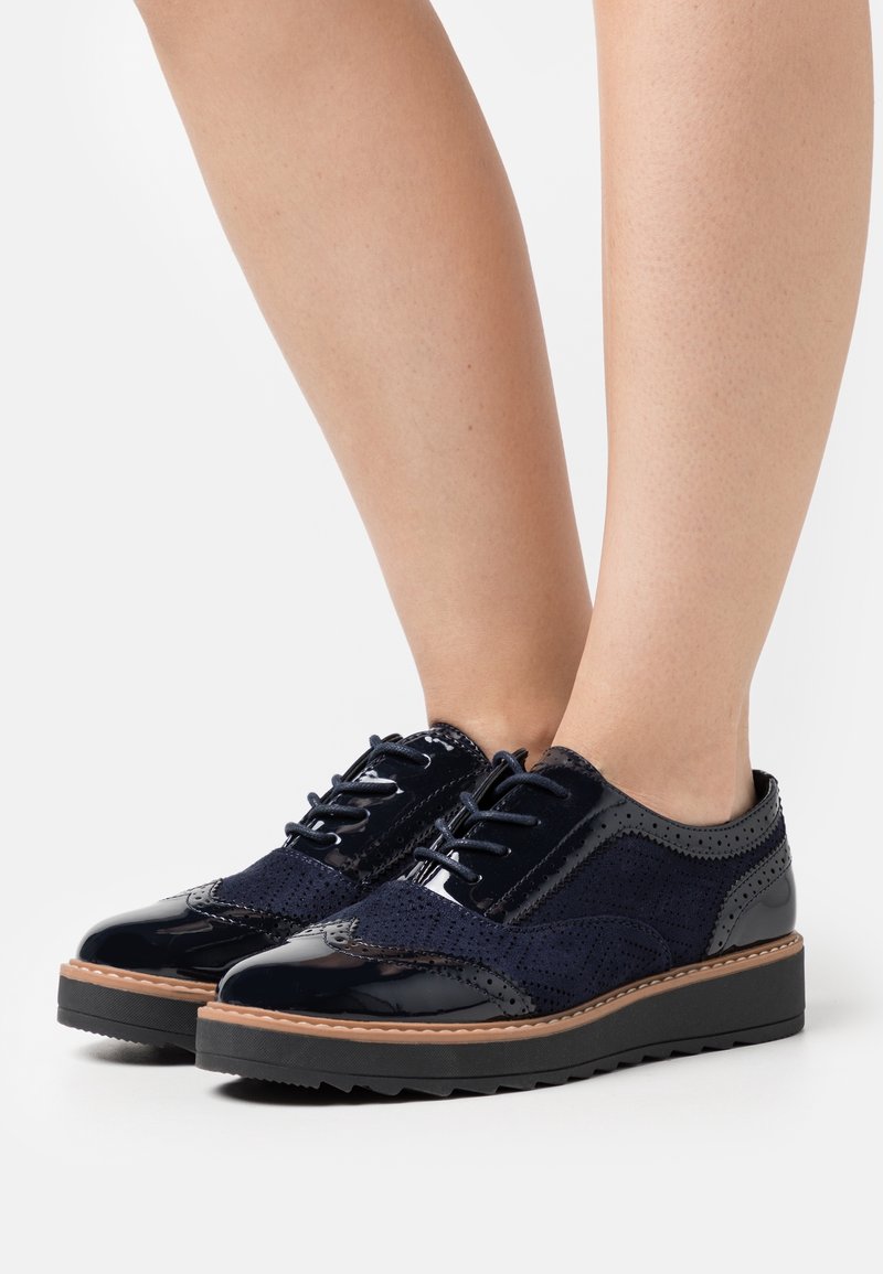 Paire de chaussures brogues bleu marine en daim perforé et cuir verni portées sur jambes nues, posées sur un fond blanc.