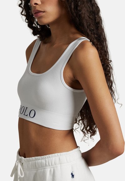 Bílý žebrovaný krátký top s širokým elastickým lemem s nápisem "POLO" v námořnické modři. Materiál je měkký s hladkou texturou a jednoduchým designem.