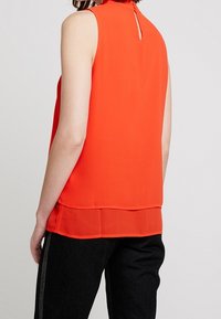 Bezramenná červená blůza s detaily ve tvaru klíčové dírky na zádech, vrstvený design a hladká textura; spojena s tmavými džíny.