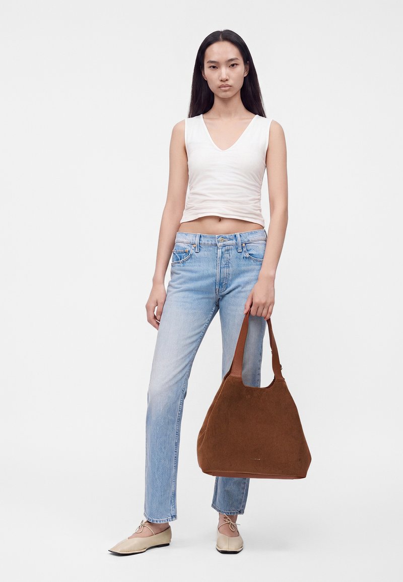 Femme portant un haut blanc sans manches et un jean bleu clair tenant un grand sac à main marron texturé sur un fond uni.