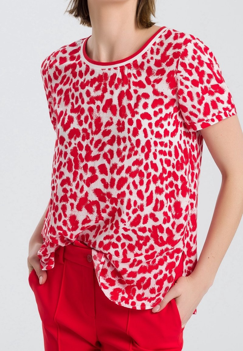 Femme portant un chemisier à manches courtes à imprimé léopard rouge et blanc, rentré dans un pantalon taille haute rouge, les mains dans les poches.