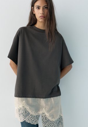 Giovane donna con lunghi capelli scuri che indossa una maglietta nera larga sopra una gonna bianca con bordi in pizzo, in piedi davanti a una parete chiara e semplice.