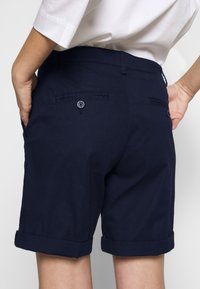 Mörkblå bomullsshorts med uppvikta fållar och bakfickor, med knappstängning och en slät yta. Enkel design och funktionella detaljer.