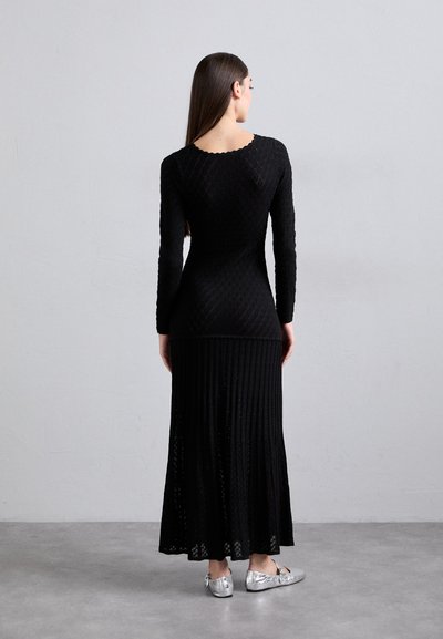 Zwarte gebreide jurk met lange mouwen, gestructureerde top en geplooide maxi-rok; gecombineerd met metallic zilveren ballerina's.