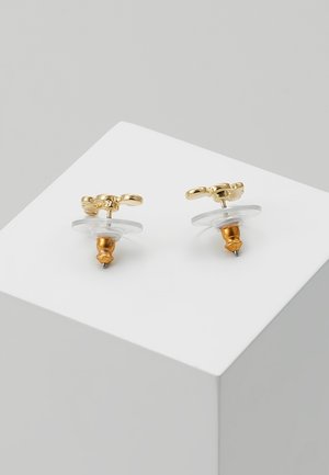Paire de petites boucles d’oreilles clous en or avec attaches protectrices transparentes présentées sur une surface blanche inclinée.