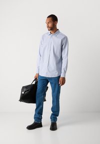 Man in een lichtblauwe overhemd met knopen, blauwe spijkerbroek en zwarte schoenen, met een grote zwarte leren tas in zijn linkerhand.